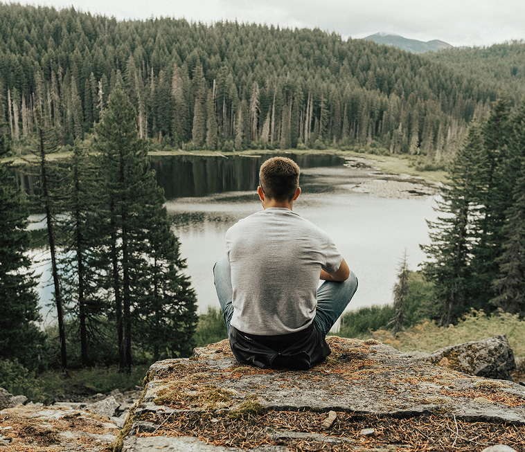 Healthy man sitting cross legged on a rock looking out over a lake with evergreen trees.